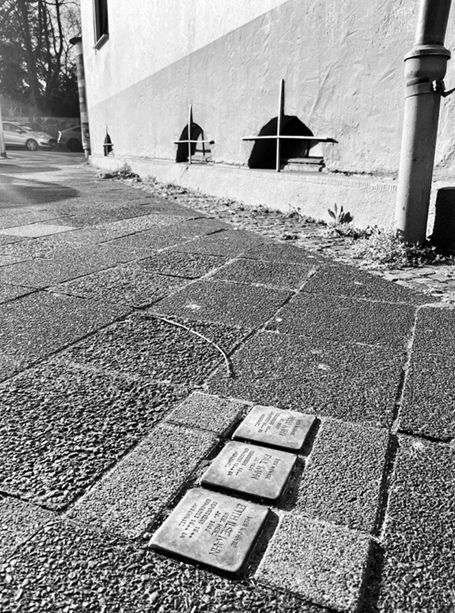 Schwarzweißfoto: drei Stolpersteine im Gehweg vor Hauswand, Pflaster und Regenrohr